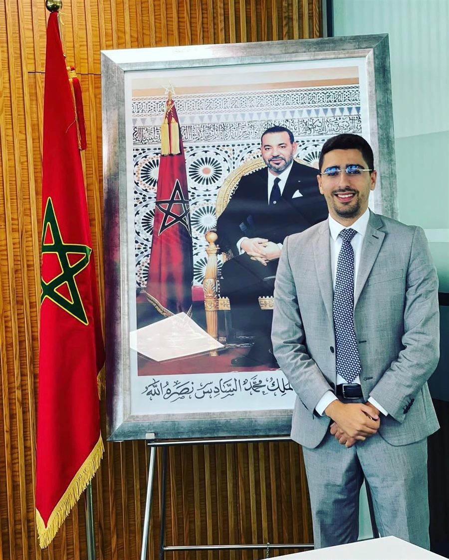 الأستاذ إسماعيل بوهمو ببدلة رسمية بجانب العلم المغربي